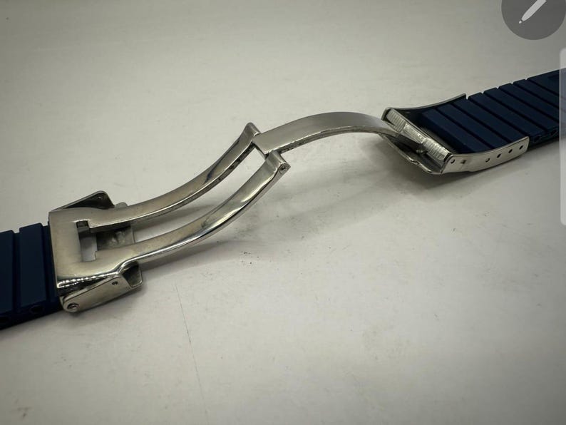 May include: A close-up of a watch strap with a silver clasp. The strap is dark blue and segmented. The clasp is open, showing the mechanism. The background is a plain, light surface.
