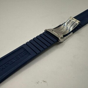 May include: A navy blue Breitling watch strap with a stainless steel clasp. The strap is made of a ribbed rubber material and features the Breitling logo. The model number 182S 22220 is visible.