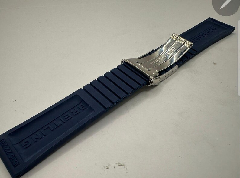 May include: A navy blue Breitling watch strap with a stainless steel clasp. The strap is made of a ribbed rubber material and features the Breitling logo. The model number 182S 22220 is visible.