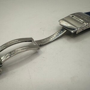 May include: A close-up of a Breitling watch clasp and navy blue watch band. The clasp is silver-toned with the Breitling logo and text. The watch band is made of a textured, dark blue material.