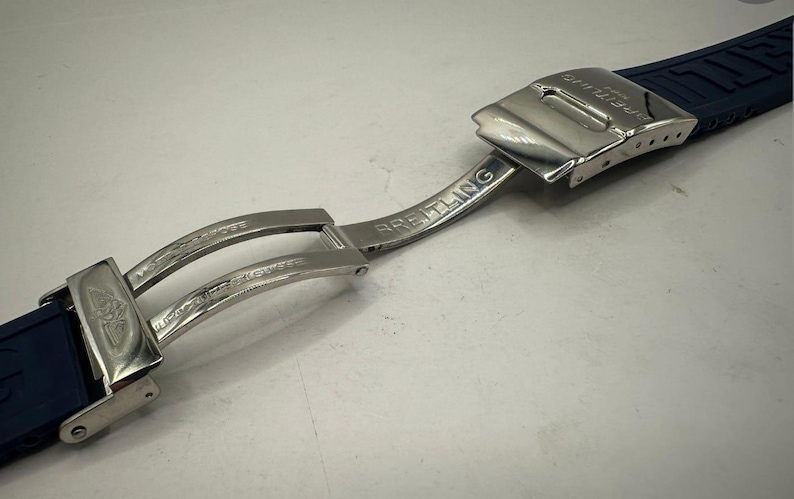 May include: A close-up of a Breitling watch clasp and navy blue watch band. The clasp is silver-toned with the Breitling logo and text. The watch band is made of a textured, dark blue material.