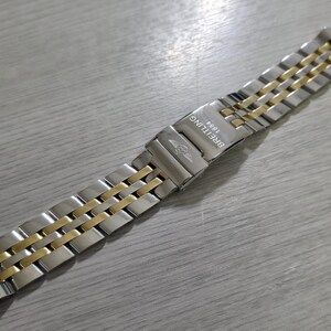 May include: A Breitling watch band with a silver and gold-tone metal design. The band is composed of rectangular links, with gold-tone links alternating between silver-tone links. The Breitling logo and "1884" are visible on the clasp.