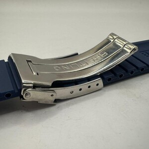 May include: A close-up of a watch strap and clasp. The strap is a dark blue rubber with a ribbed texture. The clasp is silver-colored metal with the word "BREITLING" engraved on it. The clasp is open, revealing the inner workings.