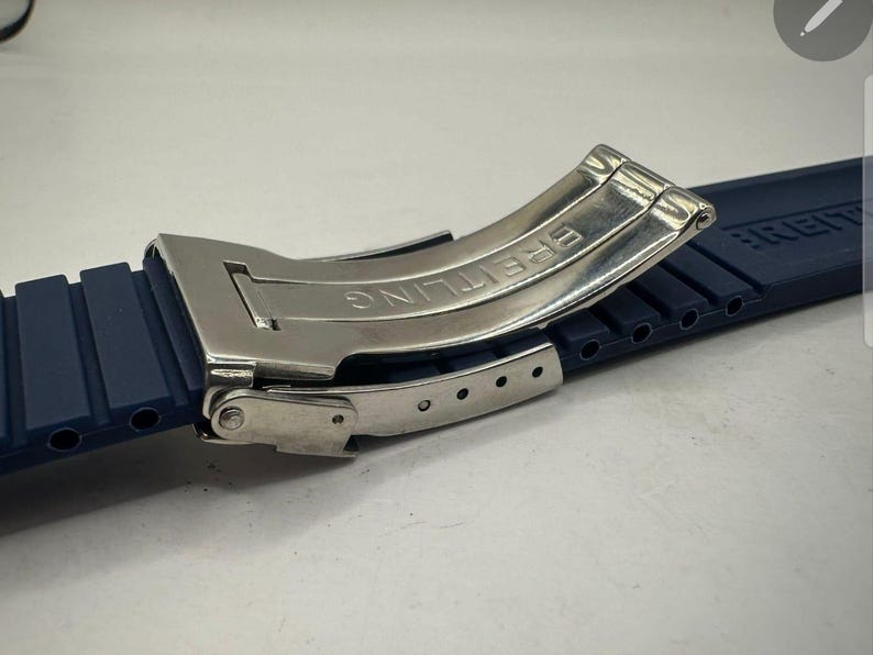 May include: A close-up of a watch strap and clasp. The strap is a dark blue rubber with a ribbed texture. The clasp is silver-colored metal with the word "BREITLING" engraved on it. The clasp is open, revealing the inner workings.