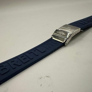 May include: A navy blue watch strap with the brand name "BREITLING" embossed along its length. The strap features a silver-colored metal clasp with the brand's logo. The strap is made of a flexible material.