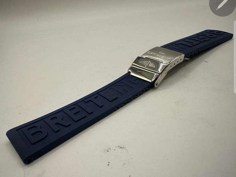 May include: A navy blue watch strap with the brand name "BREITLING" embossed along its length. The strap features a silver-colored metal clasp with the brand's logo. The strap is made of a flexible material.