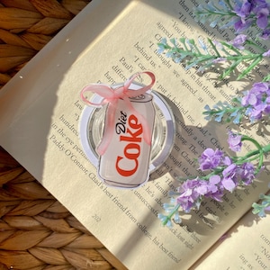 May include: A Diet Coke can-shaped sticker with a pink ribbon, placed on a clear surface. The sticker has the words "Diet Coke" in red and white. The background includes a book and purple flowers.