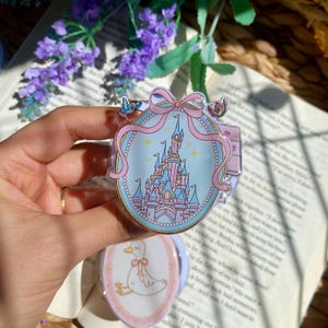 Puede incluir: Pin ovalado con un diseño de castillo rosa y azul, una cinta rosa y detalles de pájaros. También se ve un segundo pin con un diseño de pato. Los pines tienen un borde de cuentas.