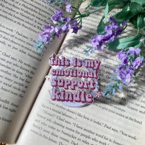 May include: A pink and white sticker with the text "this is my emotional support kindle" in a retro font. The sticker is circular with a light purple border and small flower accents. The sticker is placed on an open book with purple flowers.