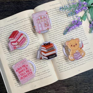 May include: A collection of decorative book-themed pins and magnets. Designs include a pink bookstore, stacks of books with a mask, a teddy bear with a knife, and a pink guest check with text. The items are set on an open book.