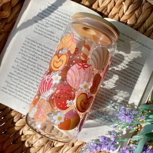 May include: A clear glass tumbler with a bamboo lid, decorated with colorful candy illustrations. The candy designs include hearts, swirls, and round shapes in shades of red, pink, orange, and white. The tumbler sits on an open book.