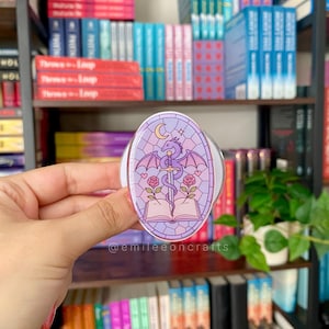 May include: An oval-shaped phone grip featuring a purple stained-glass style illustration of a dragon coiled around a sword, an open book, two pink roses, and a crescent moon. The text @emileecrafts is printed in small white letters at the bottom.