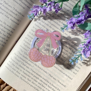 May include: A clear, round resin pin with a pink bow and two pink cherries. The pin is set against an open book with purple flowers in the background. The pin is a decorative accessory.