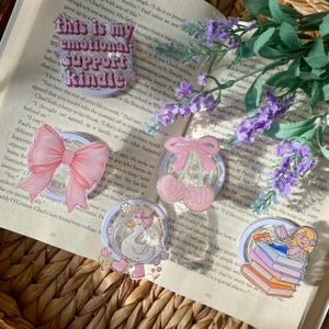 May include: A collection of clear acrylic stickers with various designs, including a pink bow, cherries, a goose wearing boots, and a girl reading. The stickers are placed on an open book with purple flowers.