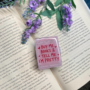 Puede incluir: Un pin rosa con forma de recibo de restaurante con el texto "BUY ME BOOKS & TELL ME I'M PRETTY" en rojo. El pin está decorado con corazones rojos y descansa sobre un libro abierto con flores moradas.