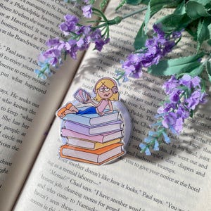 Puede incluir: Marcapáginas acrílico transparente con una chica de dibujos animados tumbada sobre una pila de libros. La chica lleva auriculares y sostiene un libro. Los libros son de varios colores, incluyendo naranja, rosa y azul. Flores moradas en el fondo.