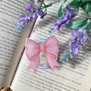 May include: A pink bow-shaped acrylic charm with a light blue backing, resting on an open book. The bow has a detailed design and is surrounded by purple flowers. A decorative accessory.