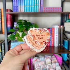 May include: A heart-shaped acrylic charm featuring a peach-colored cake illustration with white frosting, orange pumpkins, red mushrooms, and red bows. The text "Autumn Girlie" is printed in the center. The charm has a clear, glossy finish.