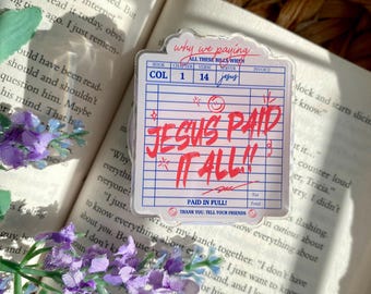 Könnte beinhalten: Ein klarer Acryl-Aufkleber im Quittungsdesign mit dem roten Text "JESUS PAID IT ALL!!". Zusätzlicher Text enthält "PAID IN FULL!" und "THANK YOU. TELL YOUR FRIENDS". Der Aufkleber liegt auf einem offenen Buch, mit lila Blumen im Vordergrund.