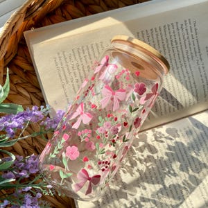 May include: A clear glass tumbler with a wooden lid, adorned with a pink floral and bow pattern. The design includes various flowers and bows in shades of pink and red. The tumbler is resting on an open book.
