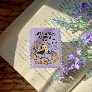 May include: A lilac-colored sticker with a cartoon duck reading a book, holding a bottle, and wearing a blanket. The sticker reads "LATE NIGHT READER CLUB" with the text "SLEEP IS OPTIONAL, BOOKS ARE HOT." The sticker is on a book page.