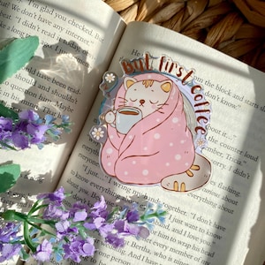May include: A clear acrylic sticker featuring a cartoon cat wrapped in a pink blanket with white polka dots, holding a coffee cup. The sticker has the text "But first coffee" in a circular design. The cat is surrounded by small white flowers.