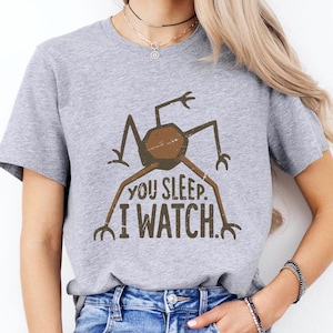 May include: A heather gray t-shirt featuring a brown graphic of a creature with the text "YOU SLEEP. I WATCH." The shirt is paired with blue denim jeans. The overall style is casual and comfortable.