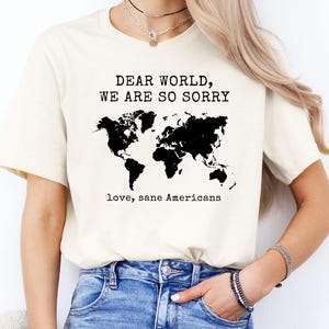 May include: Cream-colored t-shirt with the text "DEAR WORLD, WE ARE SO SORRY" above a black world map graphic. Below the map, the text reads "love, sane Americans". The shirt is paired with blue jeans.
