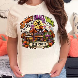 May include: A cream-colored t-shirt featuring a colorful graphic of a small town with Halloween-themed elements, including ghosts, a witch on a broomstick, pumpkins, and text that reads "SPOOKY SMALL TOWN" and "[ZIP CODE] TOWN NAME".