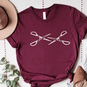May include: Burgundy t-shirt with a white graphic of two crossed scissors. The shirt is styled with a tan hat, light wash jeans, and brown flats. A beaded necklace and sprig of greenery complete the look. The shop logo "ChrissyClosett" is in the bottom left corner.