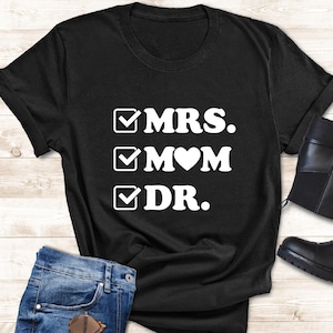 May include: Black t-shirt with the words "MRS. MOM DR." in white, with checkmarks next to each word. The shirt is paired with blue jeans, sunglasses, and black ankle boots. A small potted plant is also included.