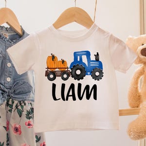 May include: A white toddler t-shirt with a blue tractor and pumpkin graphic, with the name "LIAM" in black. The shirt hangs on a wooden rack with a denim top and floral dress. A teddy bear and a hat are also on the rack.