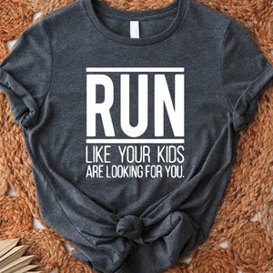 May include: Dark gray t-shirt with the white text "RUN LIKE YOUR KIDS ARE LOOKING FOR YOU." The shirt is knotted at the bottom and is displayed on a woven, circular rug.