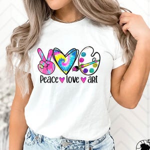 May include: White t-shirt with a colorful graphic design. The design includes a peace sign, a heart, and an artist's palette, with the words "Peace love art" below. The peace sign is pink, the heart is tie-dye, and the palette has glitter.