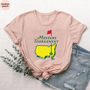 May include: A light peach-colored t-shirt featuring the text "Masters Tournament" in green, along with a yellow map of the United States with a golf flag. The shirt is paired with denim shorts.