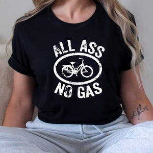 May include: Black t-shirt with the text "ALL ASS NO GAS" in white, featuring a bicycle graphic inside an oval. The shirt is worn by a person sitting down.