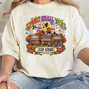 May include: A cream-colored t-shirt featuring a colorful graphic of a small town scene with pumpkins, ghosts, a witch on a broomstick, and a full moon. The text reads "SPOOKY SMALL TOWN" at the top, with "[ZIP CODE]" and "[TOWN NAME]" at the bottom.