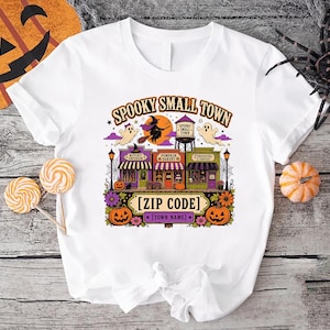 May include: A white t-shirt featuring a colorful Halloween-themed graphic of a small town with shops, a water tower, ghosts, and a witch on a broomstick. The text reads SPOOKY SMALL TOWN, [ZIP CODE], and [TOWN NAME] with pumpkins and flowers.