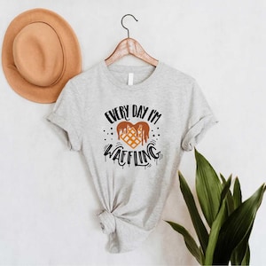 May include: A light gray t-shirt with the text "EVERY DAY I'M WAFFLING" in black and brown. The graphic features a waffle design with dripping chocolate. A tan hat hangs on the wall behind the shirt.