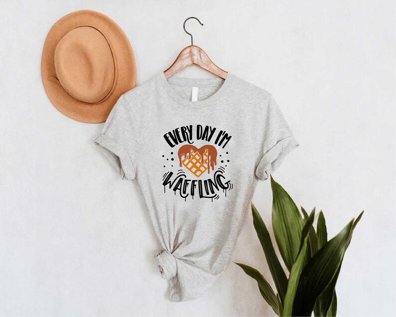 May include: A light gray t-shirt with the text "EVERY DAY I'M WAFFLING" in black and brown. The graphic features a waffle design with dripping chocolate. A tan hat hangs on the wall behind the shirt.