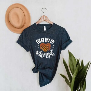 May include: A navy blue t-shirt with the text "EVERY DAY I'M WAFFLING" in white, featuring a waffle design. The shirt is hanging on a wooden hanger, with a tan hat in the background.