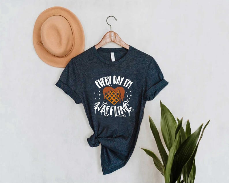 May include: A navy blue t-shirt with the text "EVERY DAY I'M WAFFLING" in white, featuring a waffle design. The shirt is hanging on a wooden hanger, with a tan hat in the background.