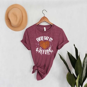 May include: Burgundy t-shirt with the text "Every Day I'm Waffling" in white, featuring a waffle heart design. The shirt is hanging on a wooden hanger. A tan hat is on the wall to the left. Green leaves are on the right.