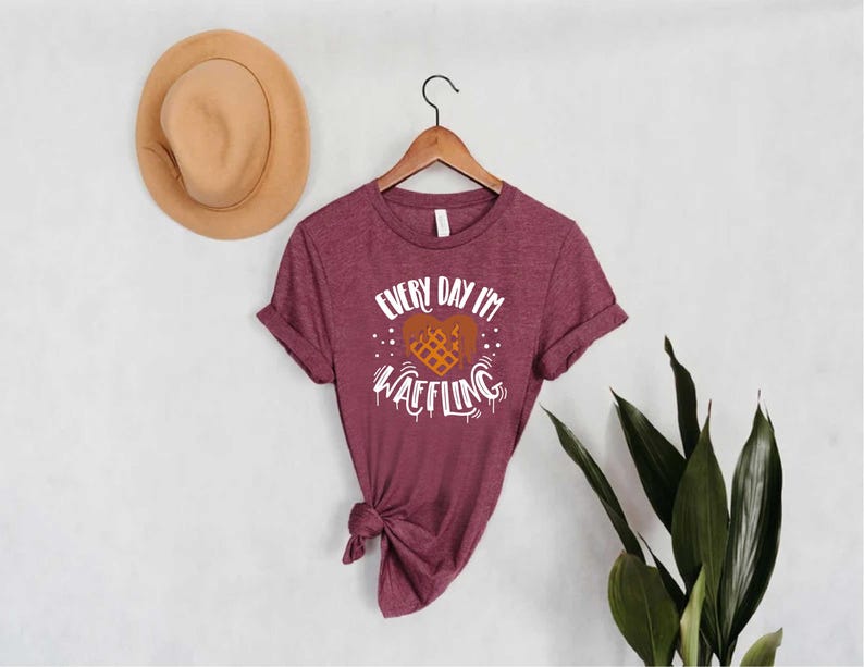 May include: Burgundy t-shirt with the text "Every Day I'm Waffling" in white, featuring a waffle heart design. The shirt is hanging on a wooden hanger. A tan hat is on the wall to the left. Green leaves are on the right.