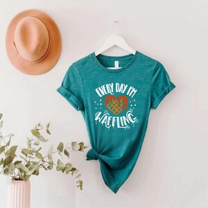 May include: Teal t-shirt with the text "Every Day I'm Waffling" and a waffle heart design. A peach-colored hat hangs above the shirt. A vase with greenery is in the lower left corner.