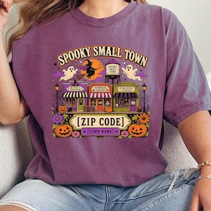 May include: A purple short-sleeve t-shirt featuring a graphic of a small town with a water tower, a witch on a broom, ghosts, and storefronts labeled Witch's Emporium, Pumpkin Bakery, and Curiosities & Follies, with text reading SPOOKY SMALL TOWN [ZIP CODE] [TOWN NAME].