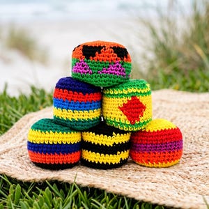 May include: A stack of six colorful, crocheted juggling balls. The balls feature various color combinations, including rainbow stripes, black and yellow stripes, and green with a red diamond. The balls are on a woven mat.