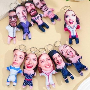 Könnte beinhalten: Eine Sammlung von Schlüsselanhängern mit personalisierten Gesichtern, die auf kleinen Plüschpuppenkörpern aufgedruckt sind. Der Text "UGLY PILLOW KEYCHAIN" ist oben zu sehen. Jeder Schlüsselanhänger hat einen Metallring.
