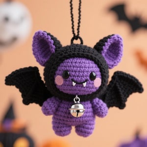 May include: A handmade crochet bat ornament in purple and black. The bat has large ears, wings, and a hood. It features a silver bell around its neck and pink cheeks. The background is a soft orange with Halloween-themed cutouts.