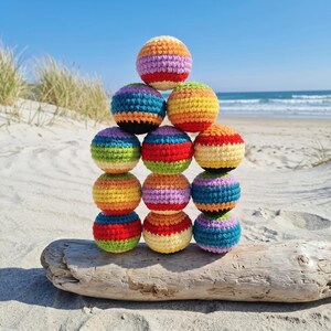 May include: A pyramid of twelve colorful, crocheted balls stacked on a piece of driftwood on a sandy beach. Each ball features a different combination of rainbow colors, including red, orange, yellow, green, blue, and purple. The background shows the ocean and blue sky.